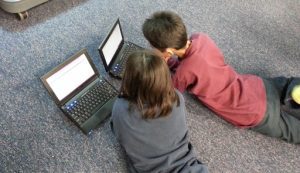 Two children lay on a carpeted floor in front of their laptops.