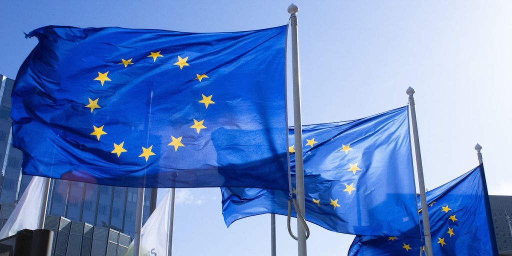 Three EU flags with a twelve yellow stars forming a circle on a blue background fly in the wind against a sunny blue sky.