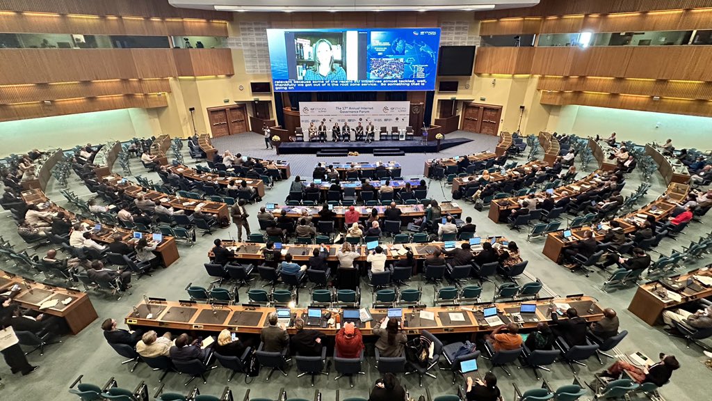 Photo of a large, filled plenary hall at IGF 2022 in Ethiopia with a panel of speakers at the front, a screen behind them, and a projection of the proceedings above them.
